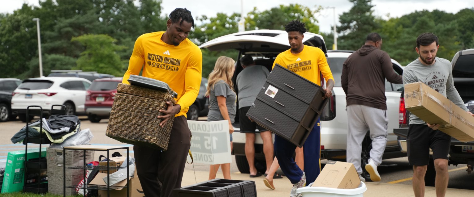Two student volunteers wearing gold WMU shirts help a new student move in to their residence hall.