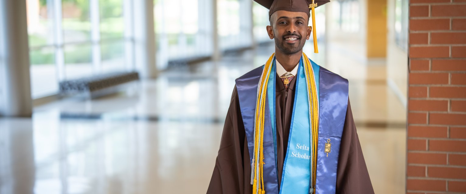 a male student poses in a brown cap and gown with a blue sash
