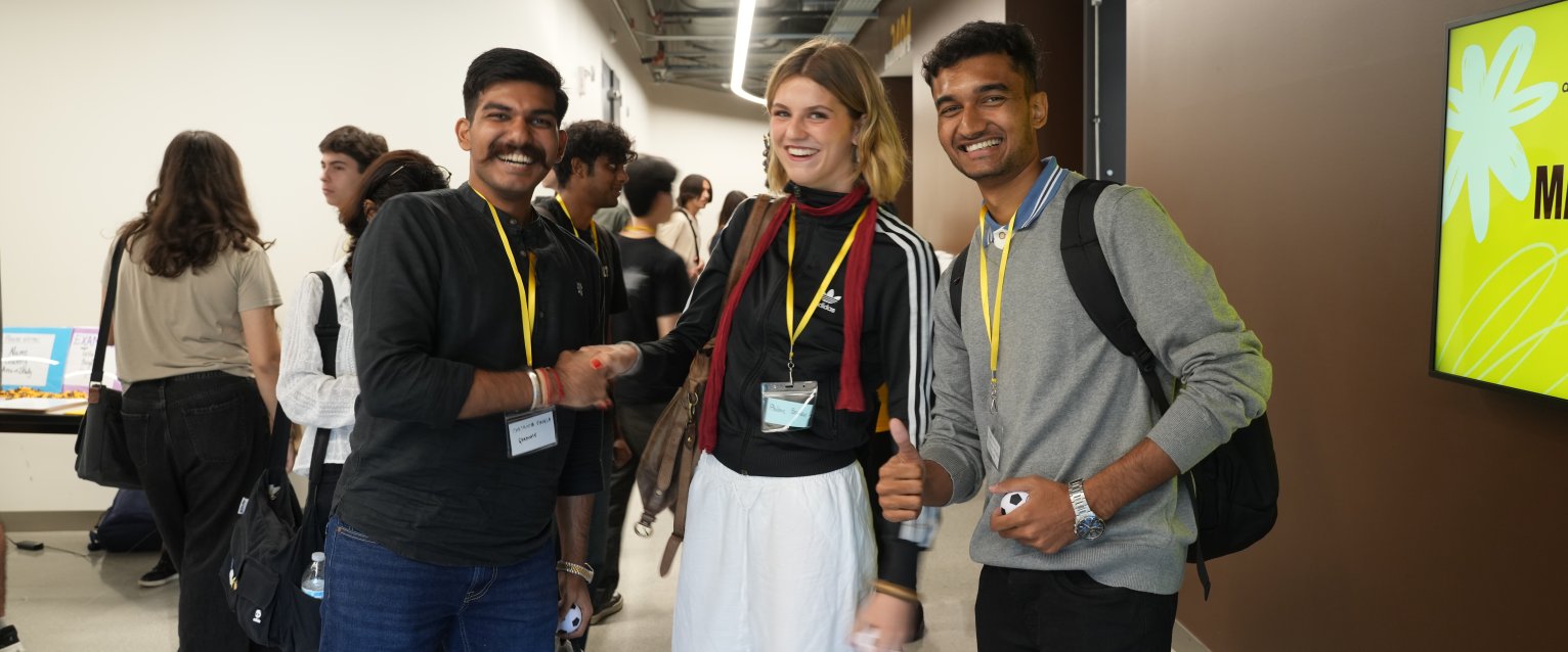 Three WMU international students smile for a picture at international student orientation in the WMU Student Center. More international students converse and browse behind them.
