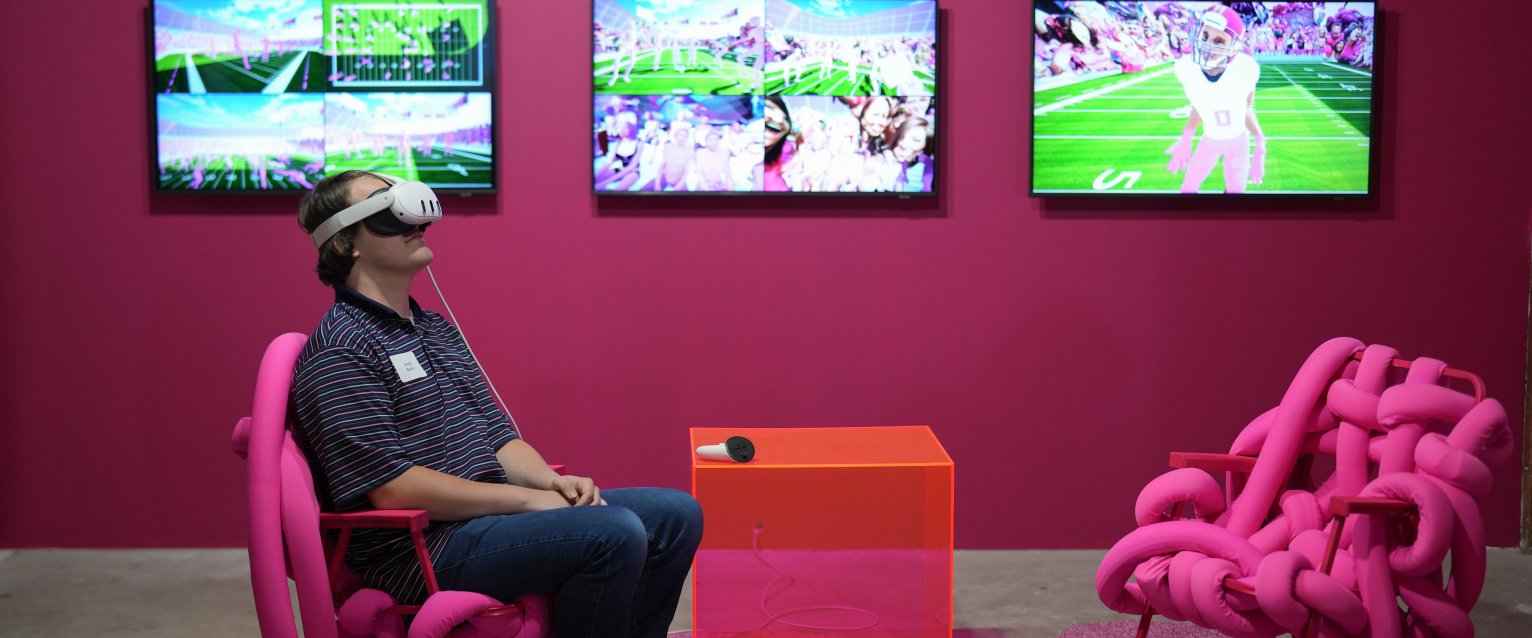 Gallery patron viewing work in a virtual reality headset, sitting in a pink room on a pink chair with screens behind them.