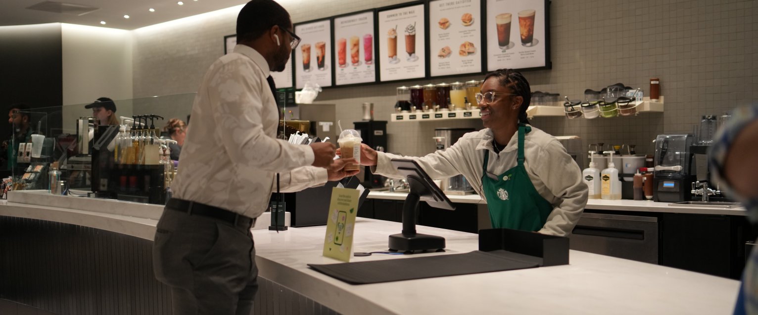 A student Starbucks employee hands a customer their coffee over the counter.