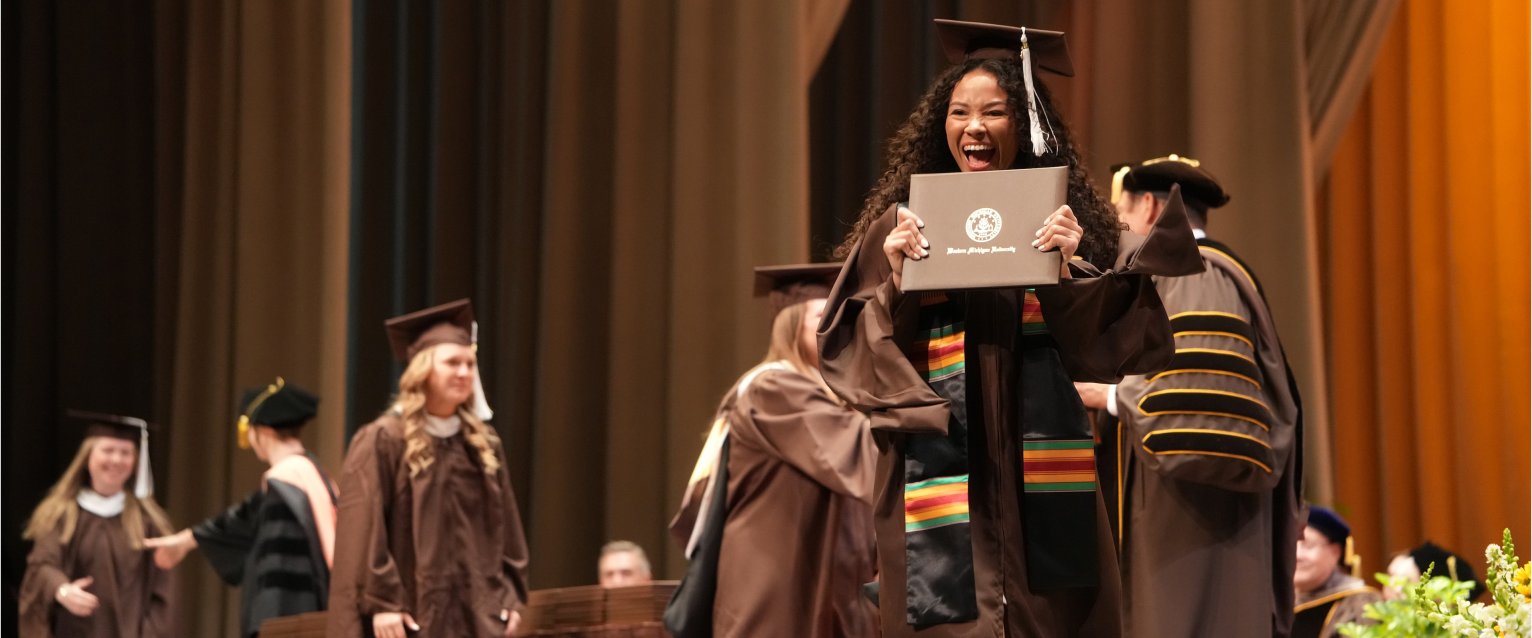 A spring 2026 Western graduate holds up her diploma with excitement as she crosses the Miller Auditorium stage.