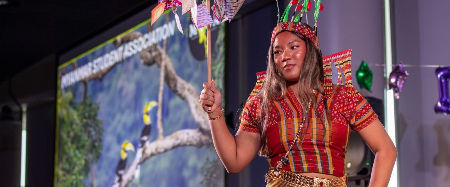 A student presents on stage during a Western Michigan University registered student organization cultural event while wearing traditional attire and holding a decorated display sign.