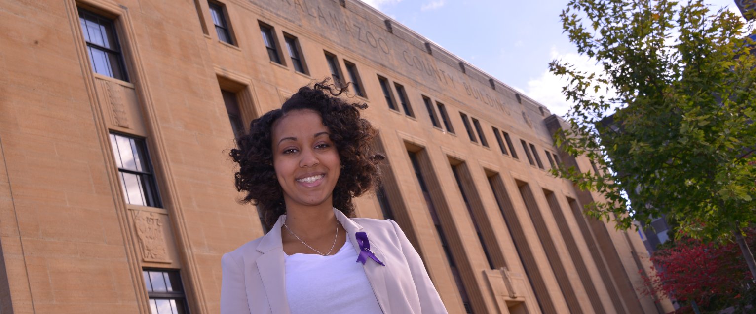Student stands outsie the Kalamazoo County Building with purple ribbon on chest. 
