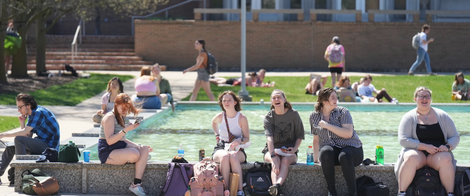 A group of students with a variety of beverages sit on an edge of Miller Fountain near Plaza Cafe.