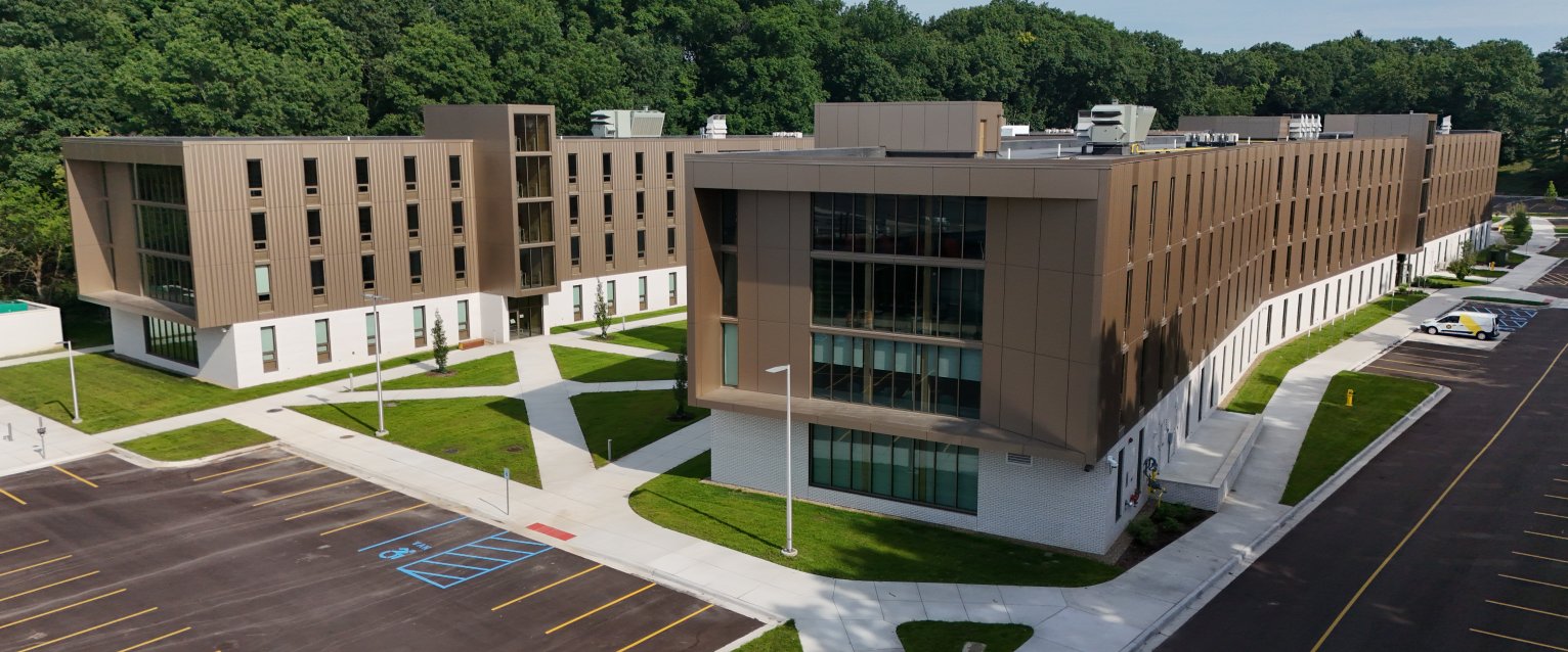 A drone shot of a modern, H-shaped residence hall with a brown exterior, large windows, and a courtyard with sidewalks cutting through it.