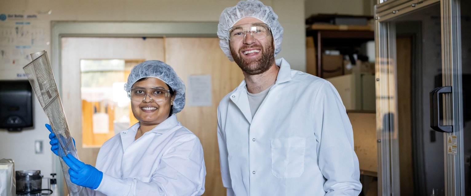 Two students in lab gear holding up a piece of clear paper with circuits on it. 