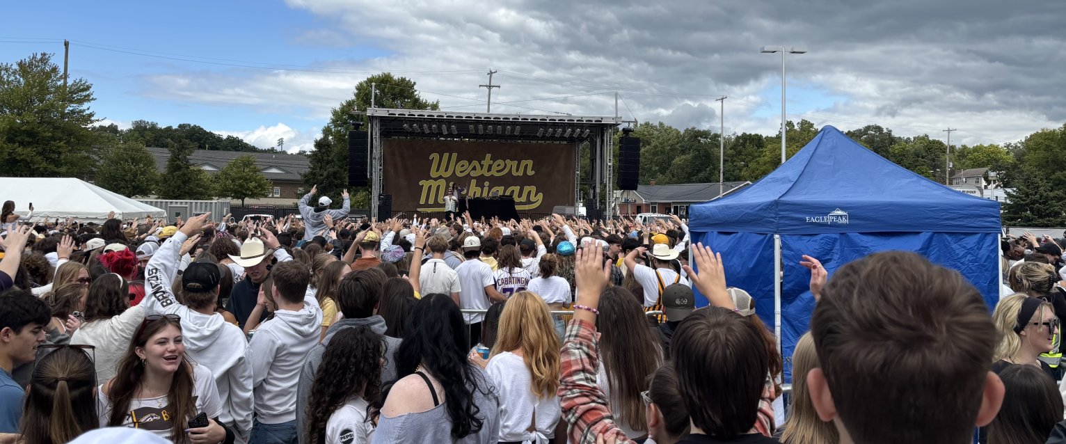 A large crowd of students stands in front of an outdoor stage with a Western Michigan backdrop, during a CAB concert performance on campus.