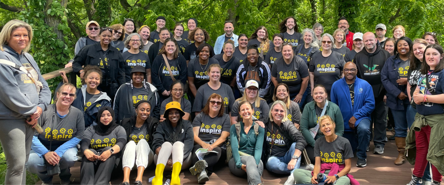 A large, diverse group of roughly 50 adults smiling and posing for a group photo on an outdoor wooden deck surrounded by lush green trees.