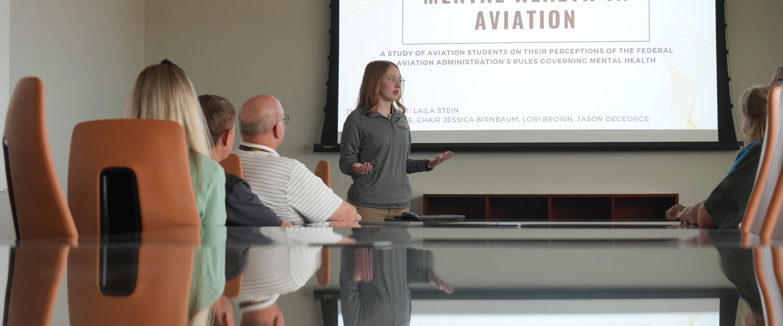 An honors student presents her research on mental health in aviation to attendees in a conference room. 