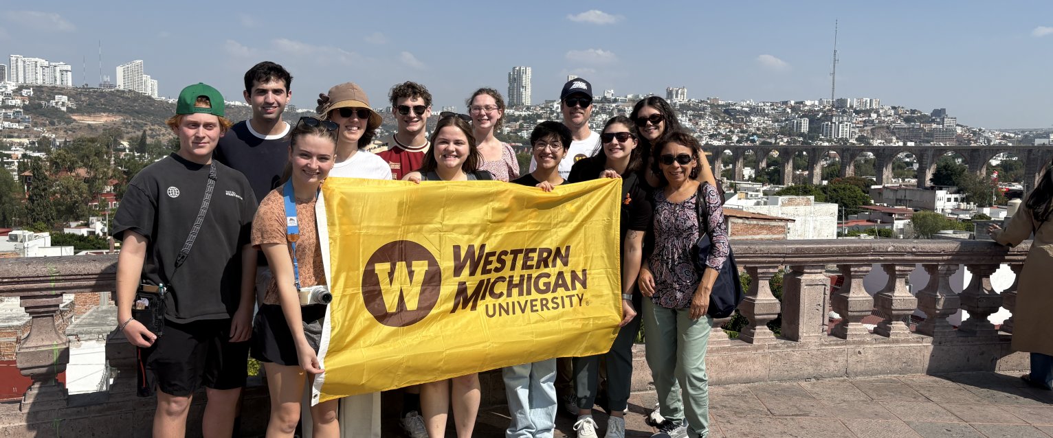 A group of honors students stands in front of a railing in Querétaro, Mexico, holding a WMU flag.