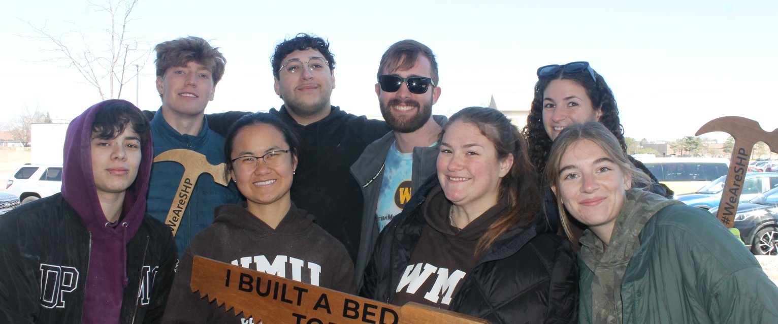 A group of honors students stands together, holding a wooden saw engraved with the words "I built a bed today."