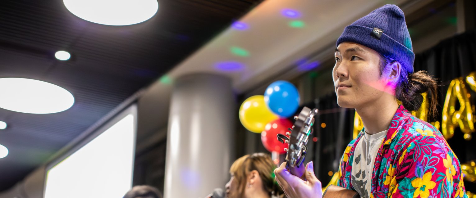 A student wearing colorful clothes plays a guitar on a small stage.