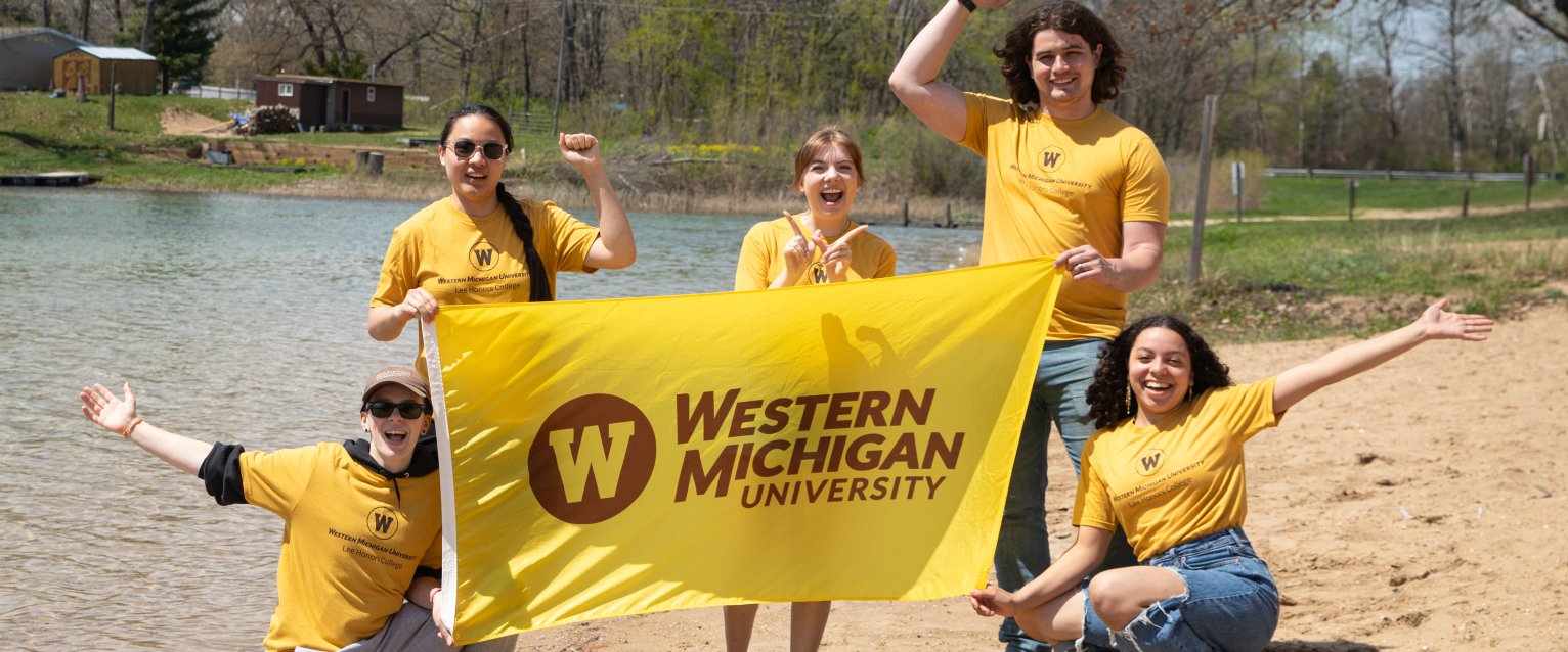 Honors students in the Idlewild course stand on the shore of a lake, holding a WMU flag.