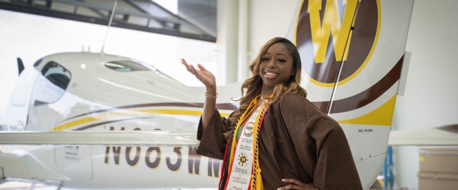 A smiling young woman in a brown graduation gown and a custom achievement stole stands inside a hangar. With one hand on her hip, she gestures proudly toward a small white, yellow, and brown airplane parked behind her, which features a large Western 'W' logo on its tail.