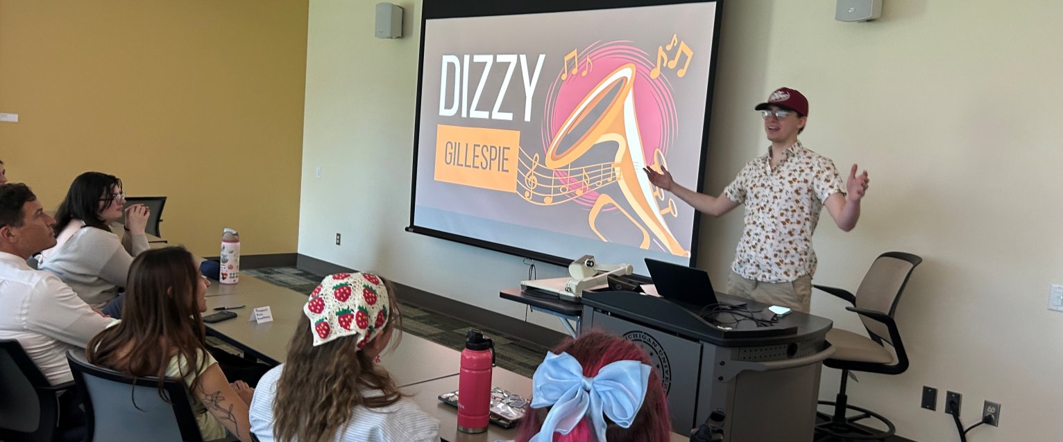 A student in a ball cap and glasses stands at the front of an honors college classroom, arms spread wide, gesturing toward the slideshow on the screen, giving a presentation on Dizzy Gillespie.