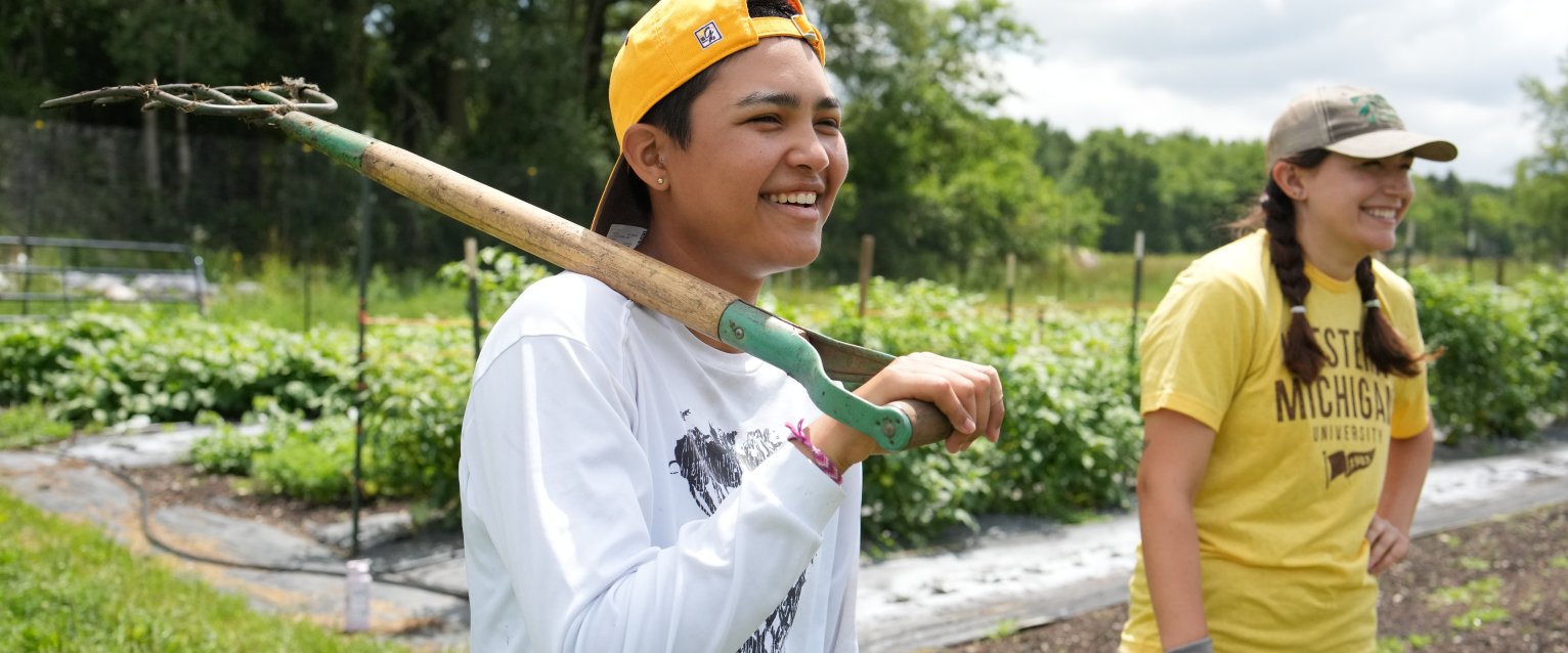a student in a white shirt holds a shovel while another stands to her right in a gold shirt in a garden