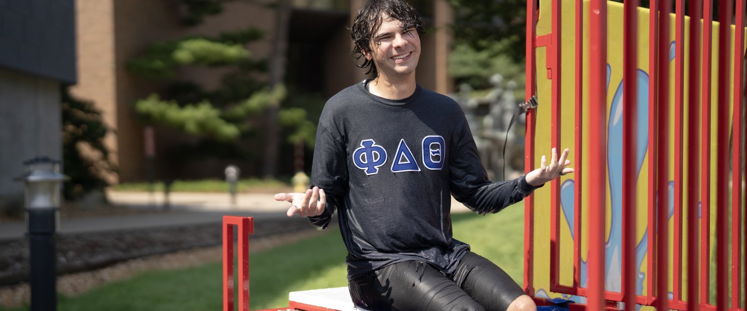 Greek WMU student wearing a fraternity shirt sits on the edge of a dunk tank after being dunked, smiling with his arms outstretched.