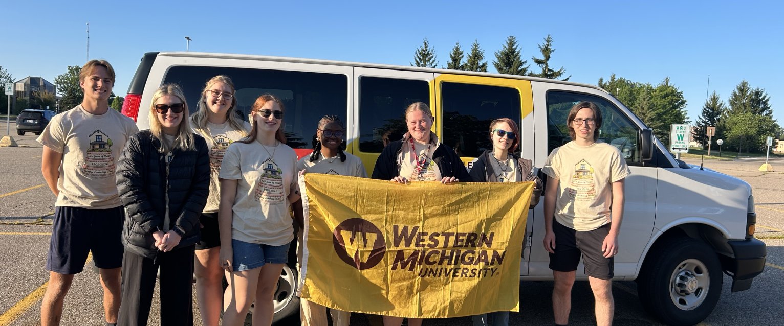 Students enrolled in the 2026 Study in the States course Walt's Pilgrimage stand next to the honors college van, holding a WMU flag, ready to depart on their trip.