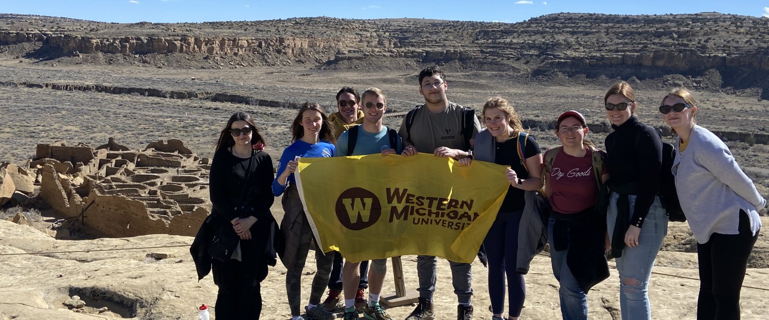 Honors students in the SIS Desert Southwest course stand near a canyon holding a WMU flag. 