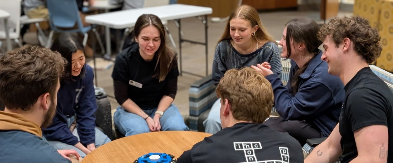 Honors college students are gathered around a round table, playing a game, in the honors college lounge. 