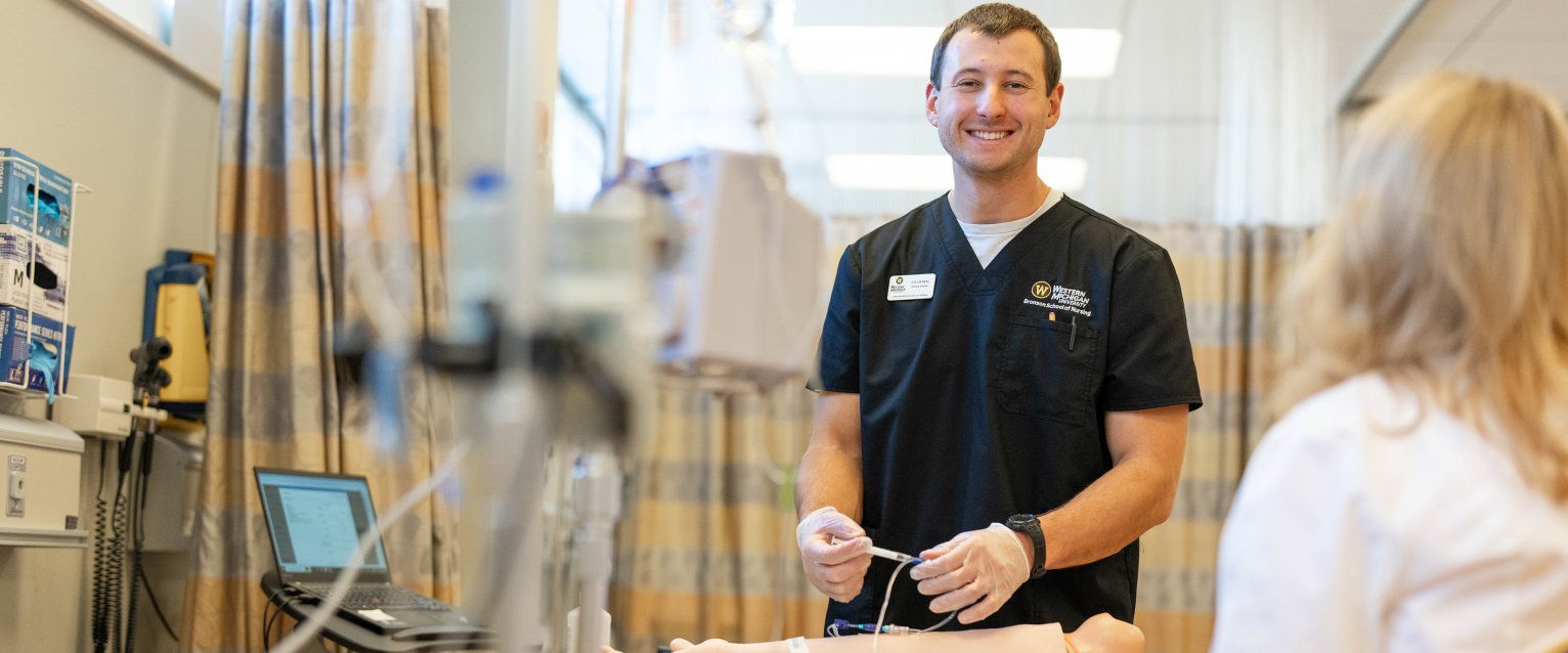 A Bronson School of Nursing student smiles at the camera while practicing injections on a patient dummy.