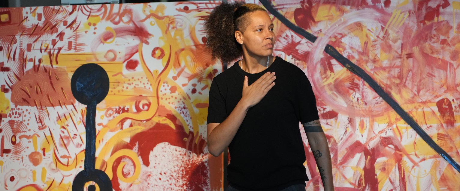 Artist Genesis Be stands with her hand over her heart in front of vibrant paintings by WMU students.