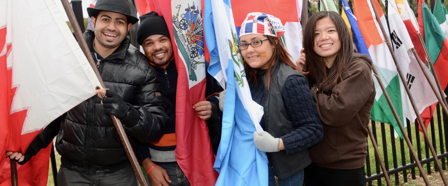 Photo of four WMU students holding and standing in front of a row of flags from several countries.