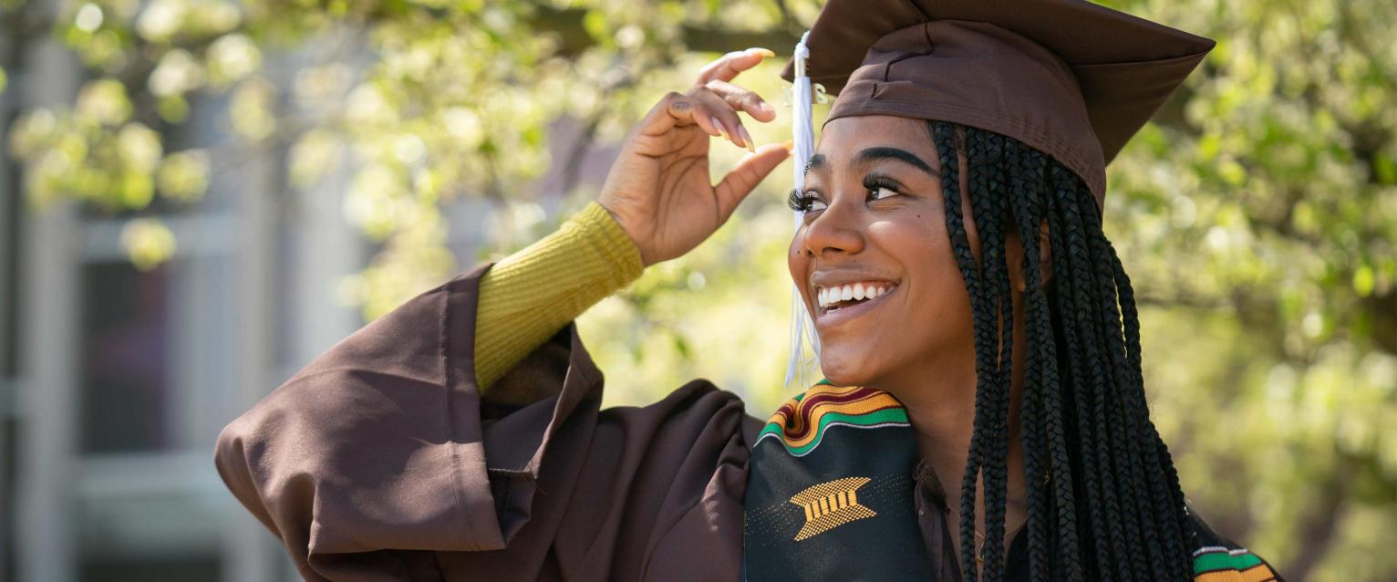 A woman wearing graduation regalia smiles outside on campus.
