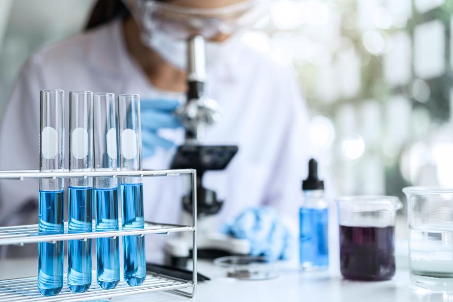 Scientific test tubes filled with blue liquid stand up in the foreground while a scientist looks into a microscope in the background.