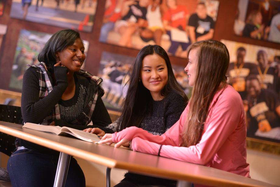 Three students sitting at a table, with a book open, facing each other and smiling.