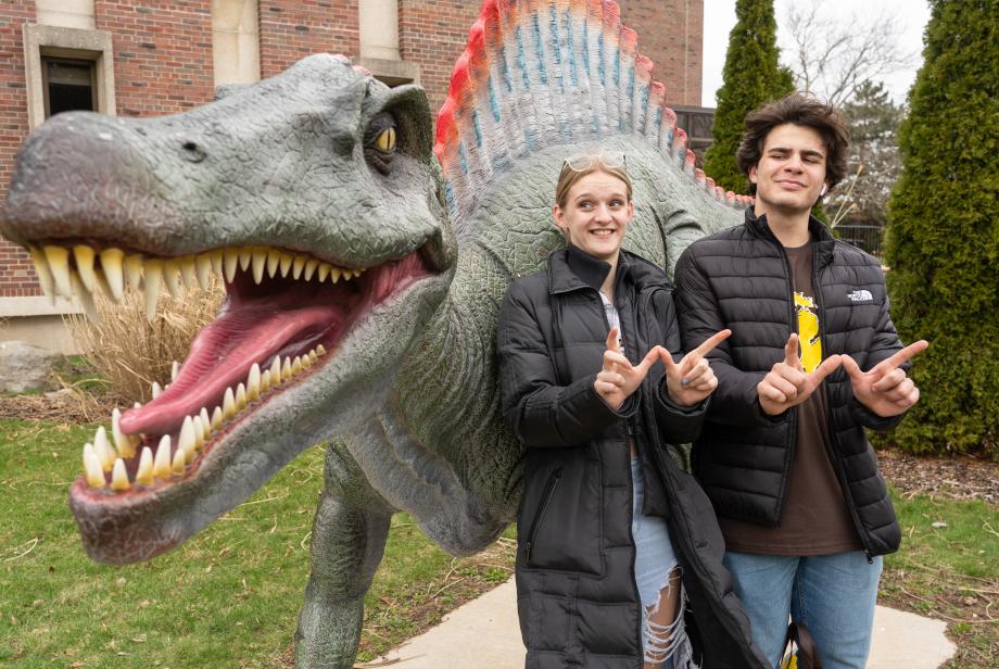 Two students facing the camera while making the W sign and standing in front of a spinosaurus statue in Rood Hall Dinosaur Park.