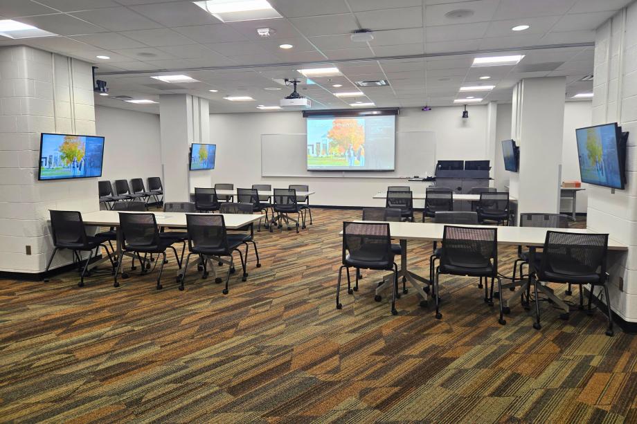 View from back of Multipurpose Room 1070 in Waldo Library showing large screen at front of room and four monitors mounted on columns with table and chairs below.