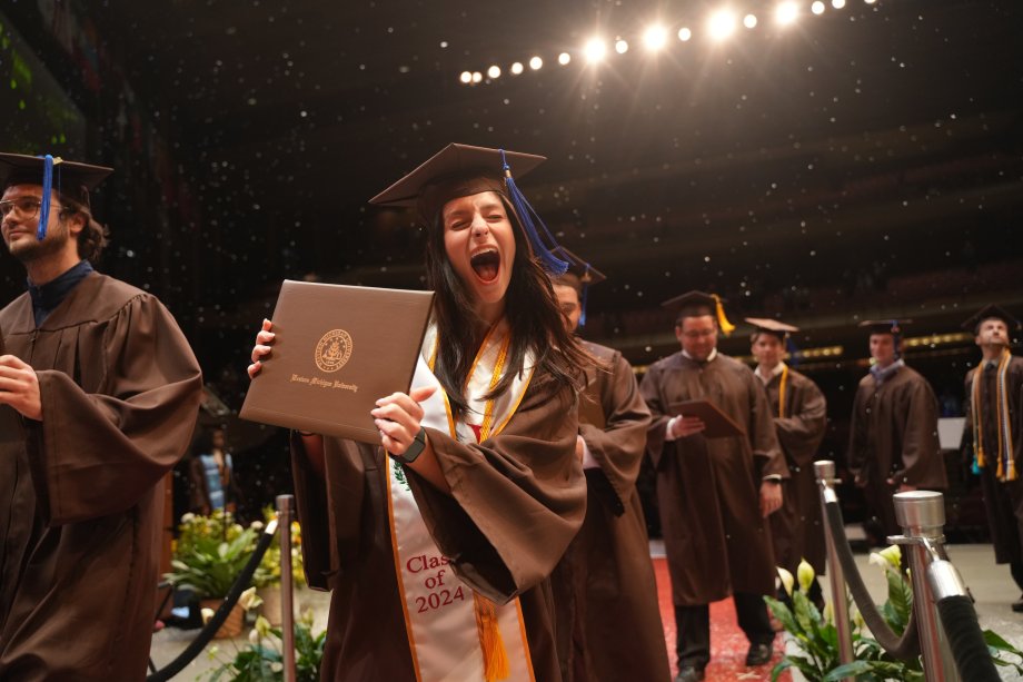 A WMU graduate wearing her graduation cap and gown proudly holds up her diploma.