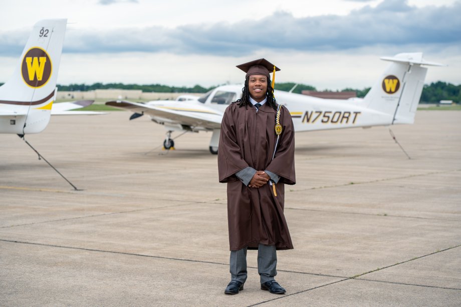 Raymond Miller stands on the tarmac at the College of Aviation while wearing his graduation cap and gown.