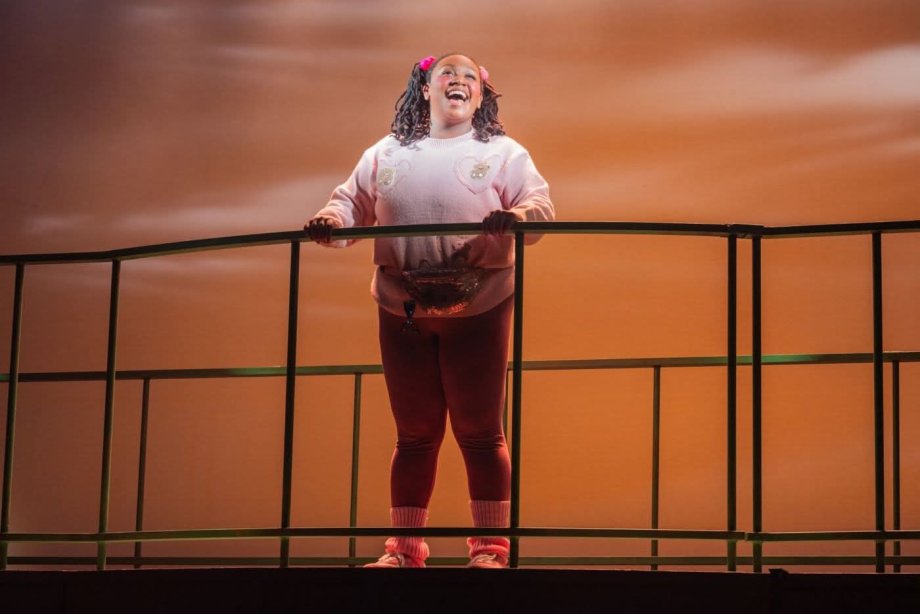 Black woman on stage, wearing a pink sweatshirt and pigtails, singing andsmiling.