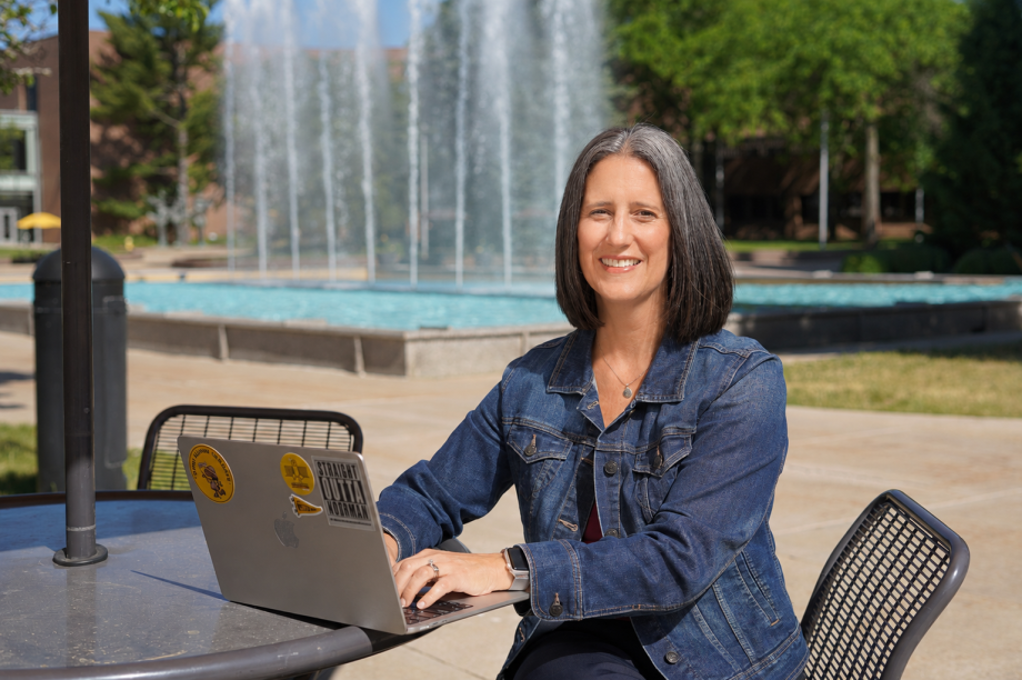 Amy Coughenour smiles while working on a laptop at an outdoor table in front of Miller fountain.