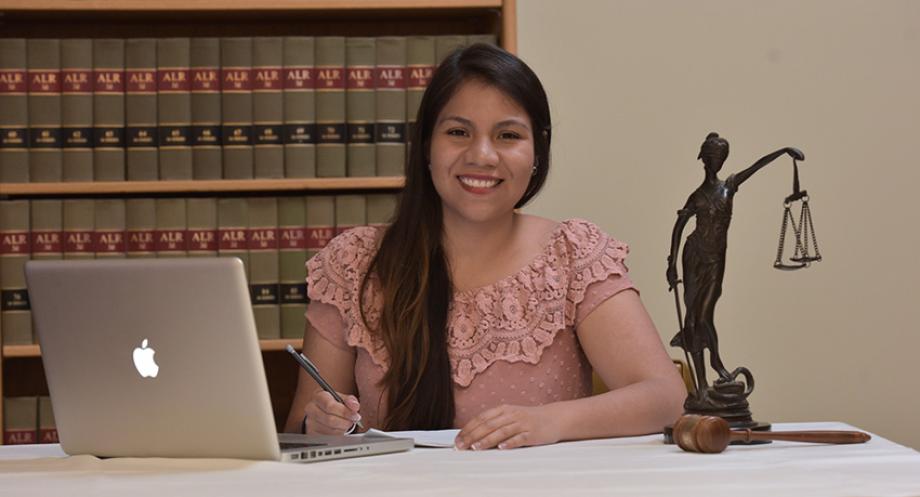 Student sitting at desk with laptop, Lady Justice statue and judges gavel in background.