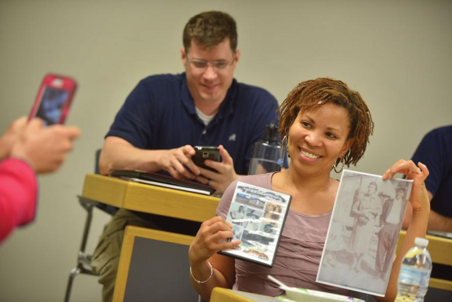 A student holds up pictures while someone takes a photo with a cell phone.