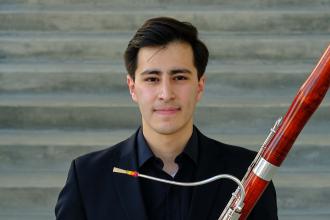 A smiling man in a dark suit, holding a bassoon.