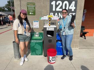 Students stand next to recycling and trash bins.