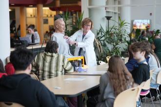 A professor in a lab coat points to a mannequin.