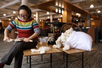 Person wearing colorful and striped sweater sits on a bench working with a spinning tool. Next to her are piles of unspun wool. 