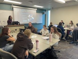 Professor William McQuitty lectures in a classroom while students listen at nearby tables.
