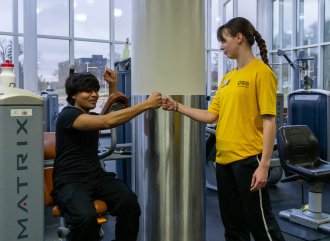 Female personal trainer in a yellow staff t-shirt fist bumping with a male client who is sitting and working out on a machine. 