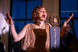 Maya Narayan sings into a microphone labeled "Gatsby" during a theatre performance.