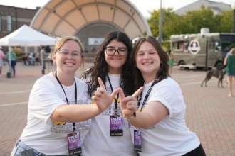 Three smiling young women dressed in Western Michigan University event staff t-shirts and lanyards, showing the WMU "W" hand sign. Behind them, an outdoor amphitheater stage, a food truck, and event tents are visible, along with other attendees.