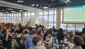 A crowd of people sitting around tables. They're listening to a presentation where the speaker has a powerpoint on the screen. 