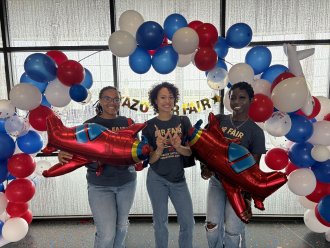 Three students hold balloon airplanes in front of a balloon arch at a volunteer event.