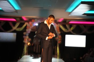 A runway model in a dark pinstripe suit with a high leg slit, adjusting his cuff.